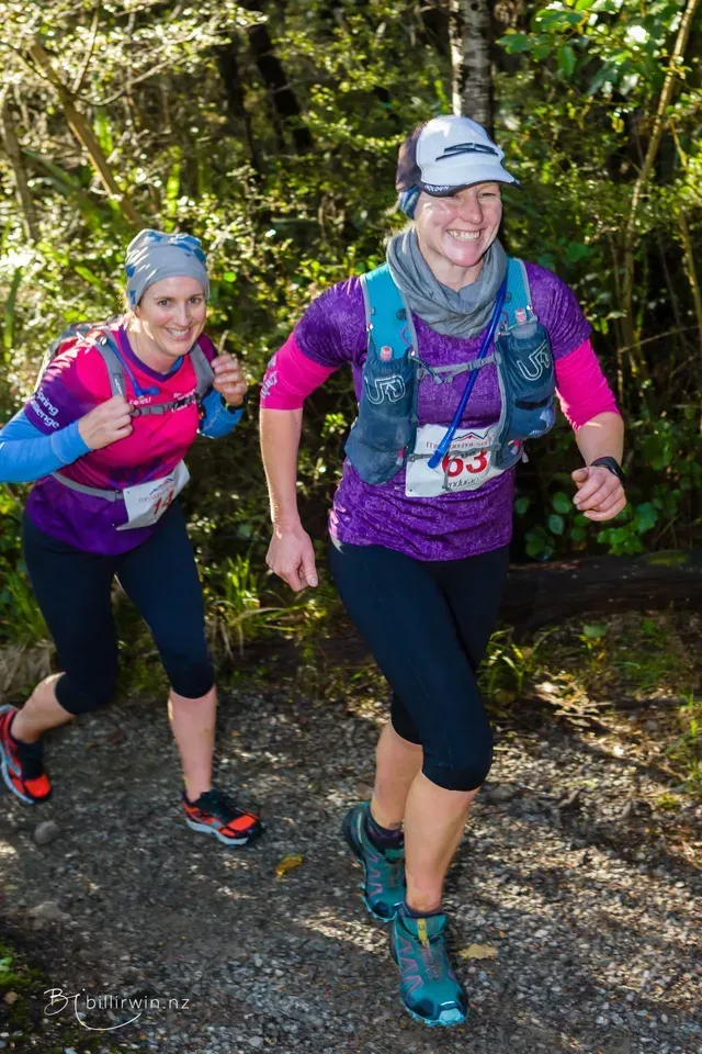Two women are running on a trail in the woods.