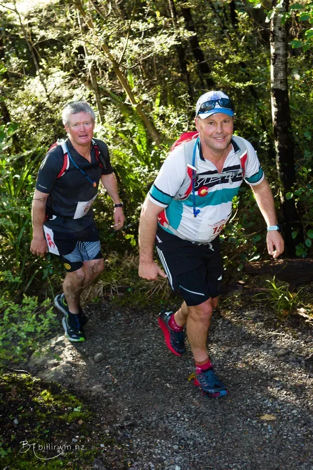 Two men are running down a trail in the woods.