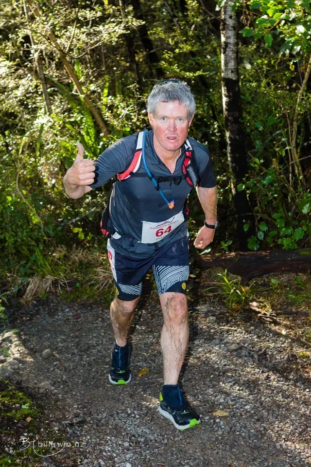 A man is running on a trail in the woods and giving a thumbs up.