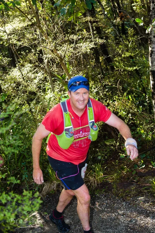 A man in a red shirt and green vest is running through the woods.