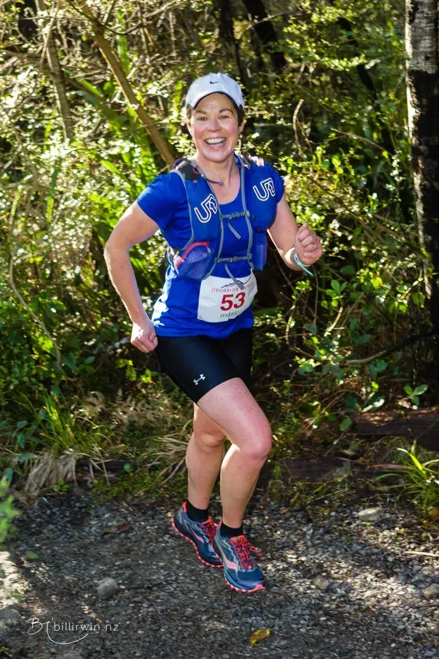 A woman in a blue shirt and black shorts is running in the woods.