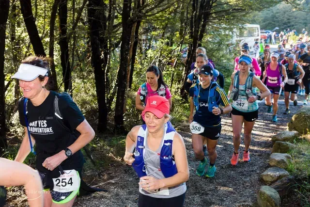 A group of women are running down a trail in the woods.