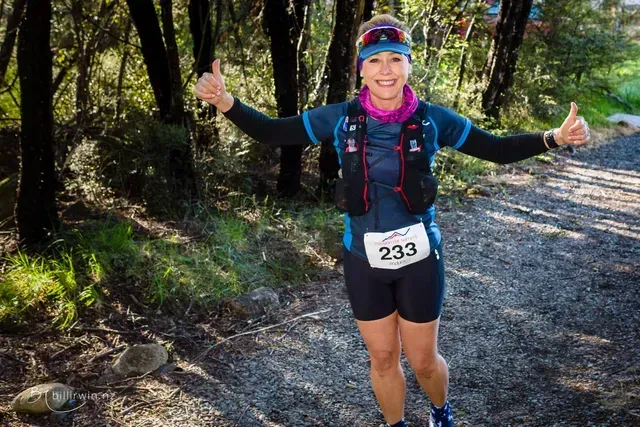 A woman is standing on a dirt path in the woods giving a thumbs up.