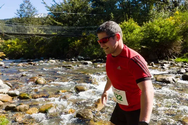 A man wearing sunglasses and a red shirt is crossing a river.