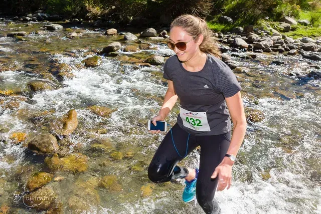 A woman is crossing a river while holding a cell phone.