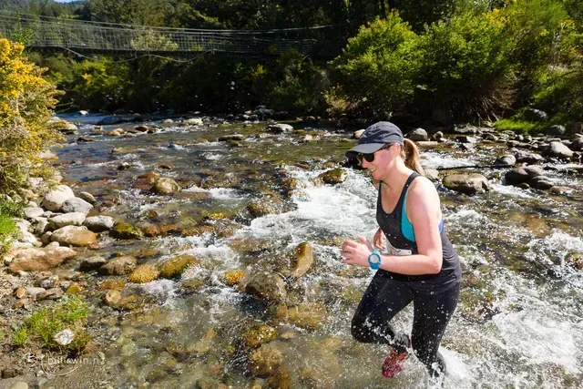 A woman is walking through a river with a bridge in the background.