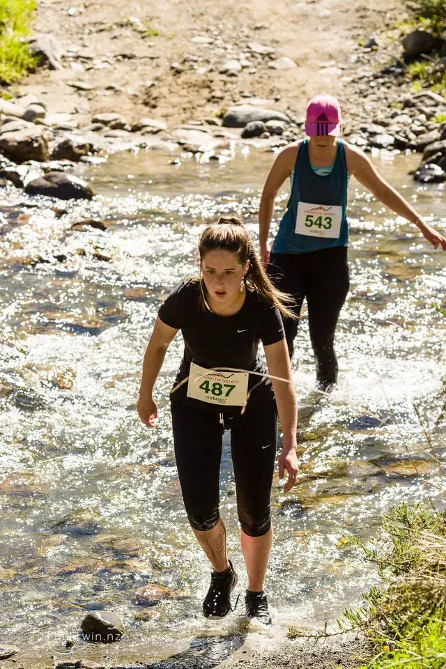 Two women are crossing a stream in a mud run.