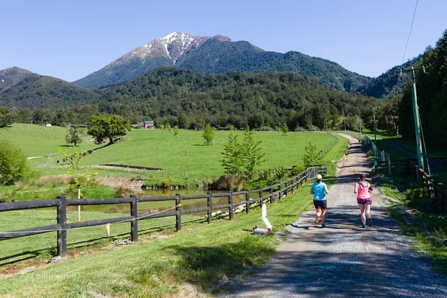 Two people are walking down a dirt road with a mountain in the background.