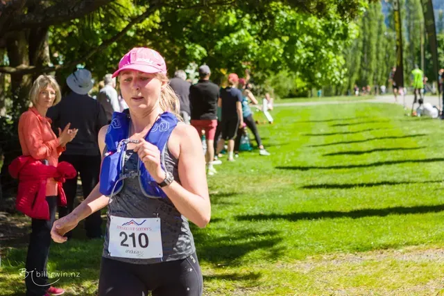 A woman is running in a park with a number 210 on her shirt.