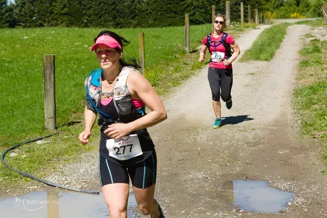 Two women are running on a dirt road and one has the number 277 on her shirt.