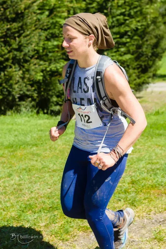 A woman wearing a bandana and a backpack is running on a grassy hill.