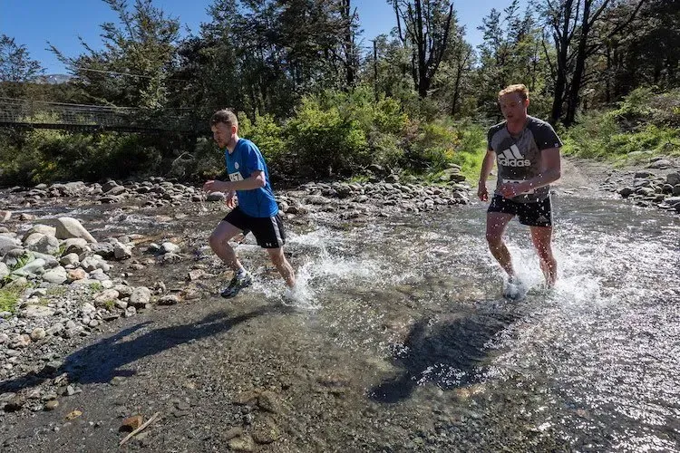 Two men are running through a stream in the woods.