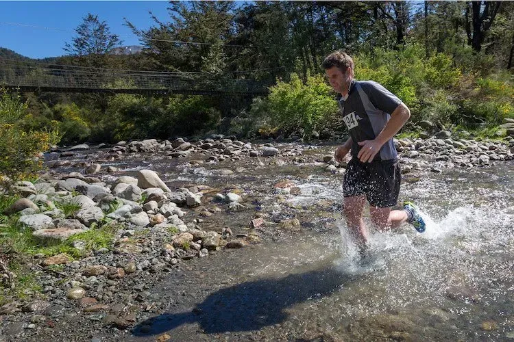 A man is running through a stream wearing a shirt that says asics