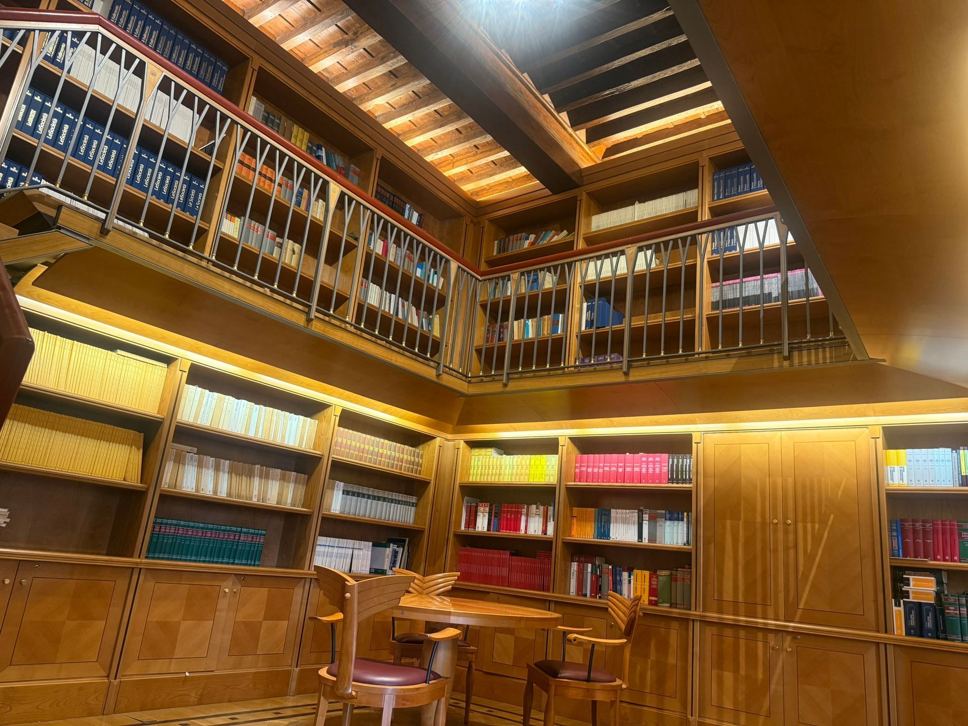 Biblioteca in legno con scaffali a due piani pieni di libri, tavolino, sedie e illuminazione calda.