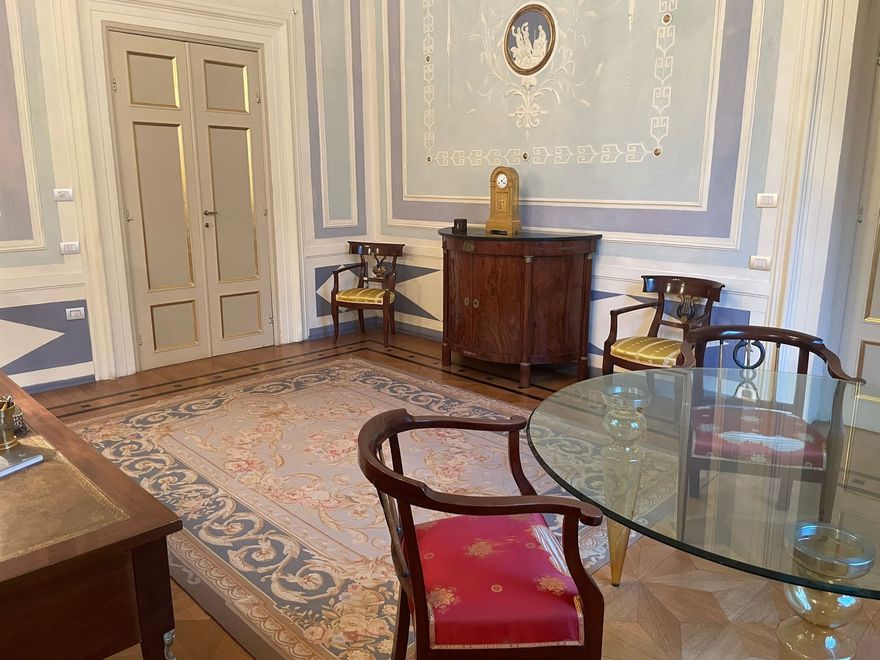 Un ufficio con una grande scrivania in legno, un divano rosso e una porta ad arco che conduce alla biblioteca.