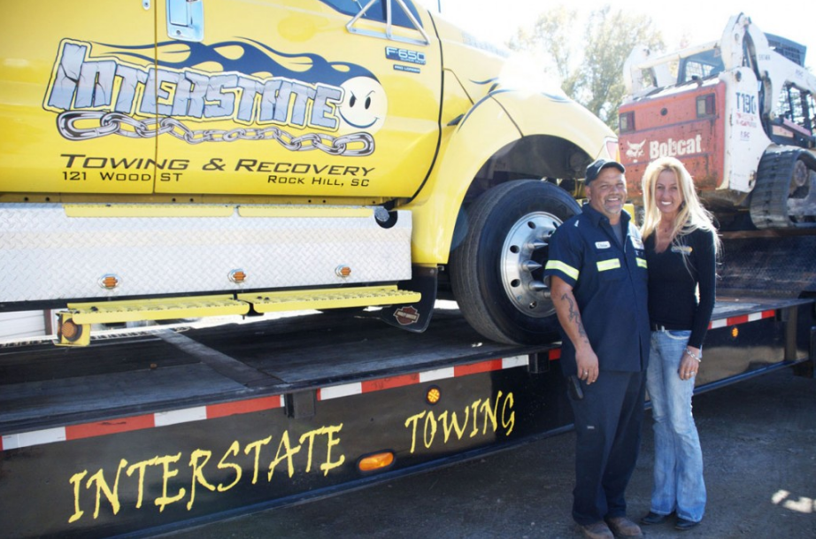 A man and a woman are standing in front of an interstate towing truck