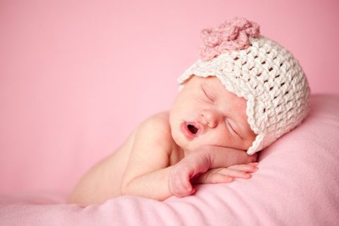 Color photo of a beautiful newborn baby girl wearing a crocheted hat while sleeping peacefully on a pink background.
