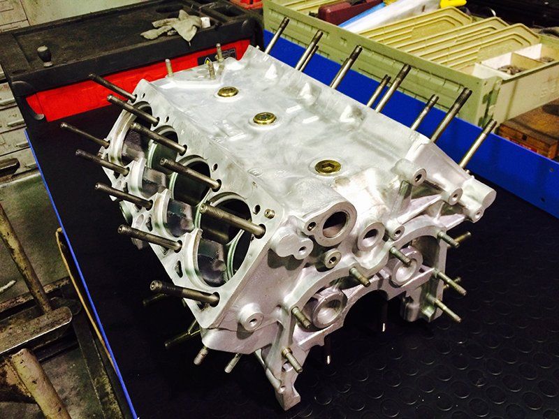 Un blocco motore in alluminio pulito e smontato, con le canne dei cilindri e i prigionieri di fissaggio a vista, appoggiato su un banco da officina.