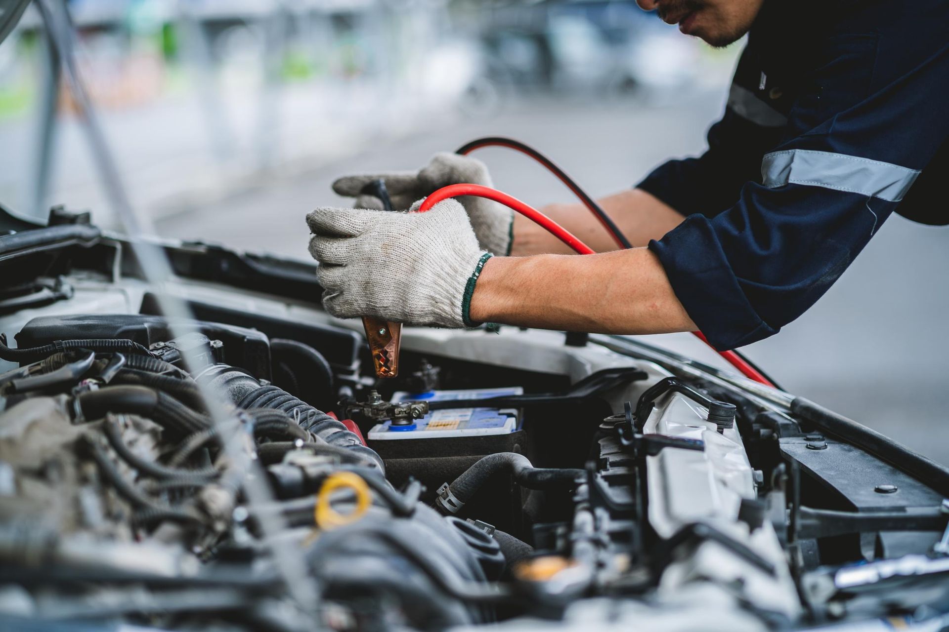 A person wearing work gloves using red jumper cables to jump-start a car battery .