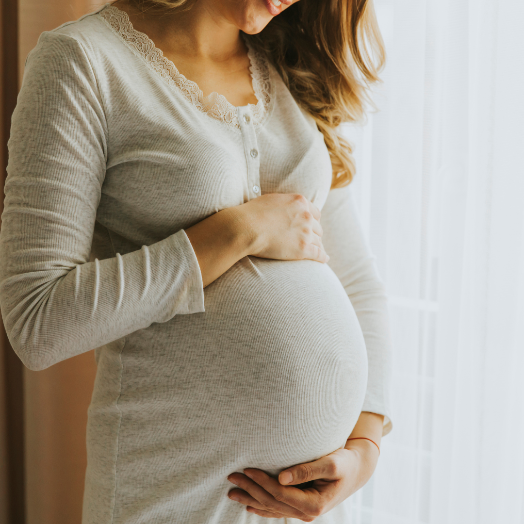 A pregnant woman is standing in front of a window holding her belly.