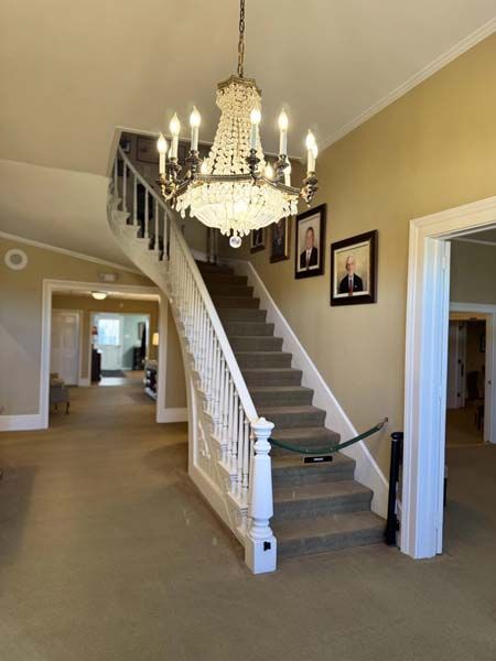 Grand staircase with ornate chandelier, portraits, and light-colored carpet and walls.