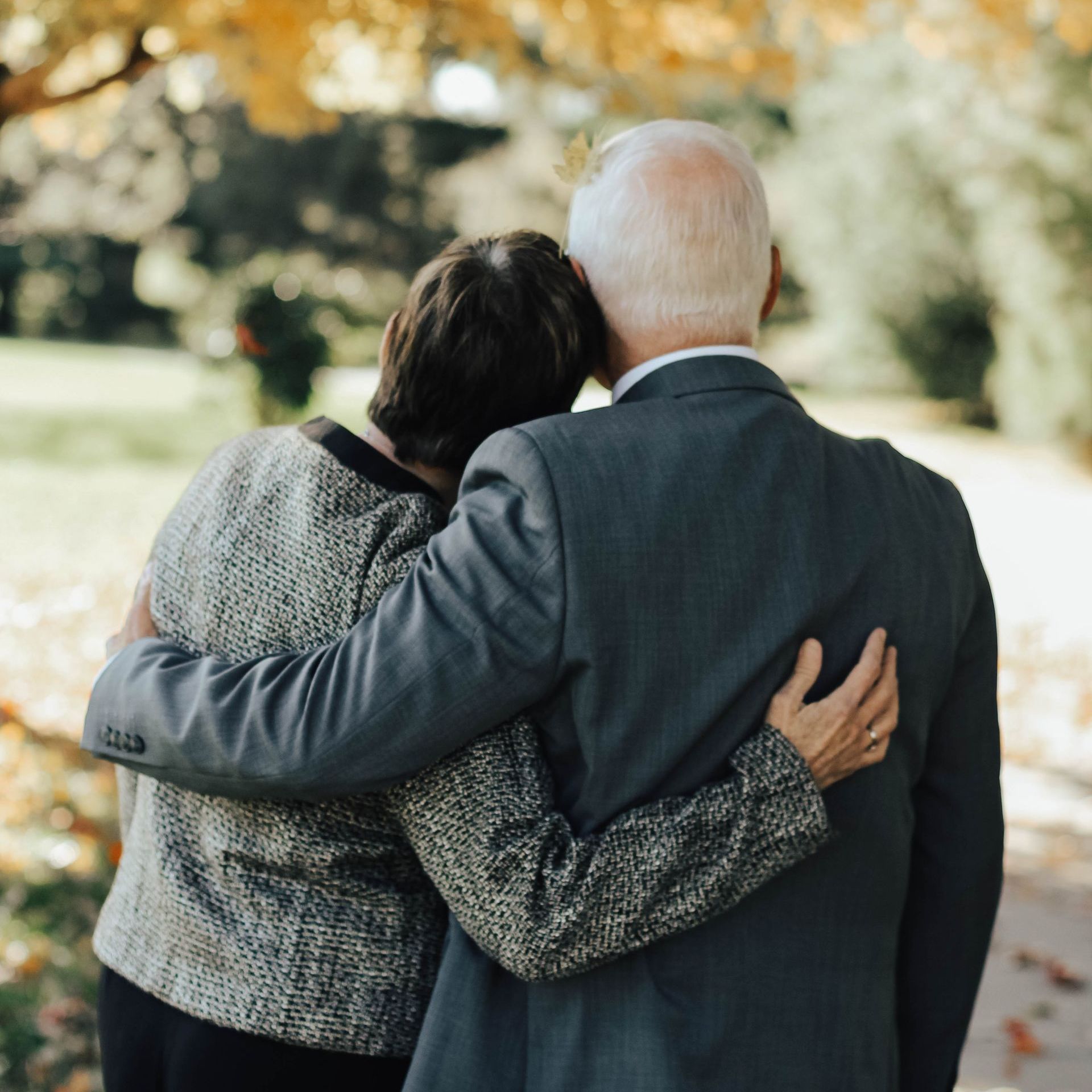 An older couple with arms around each other, walking outdoors near fall foliage.