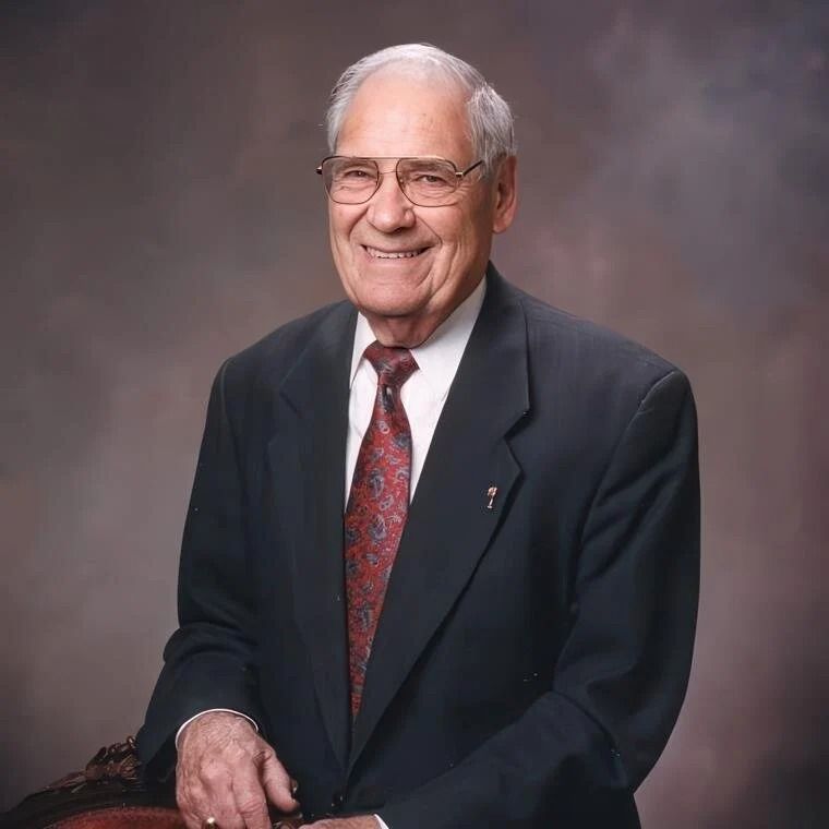 Older man in suit and tie, smiling, seated with a neutral background.