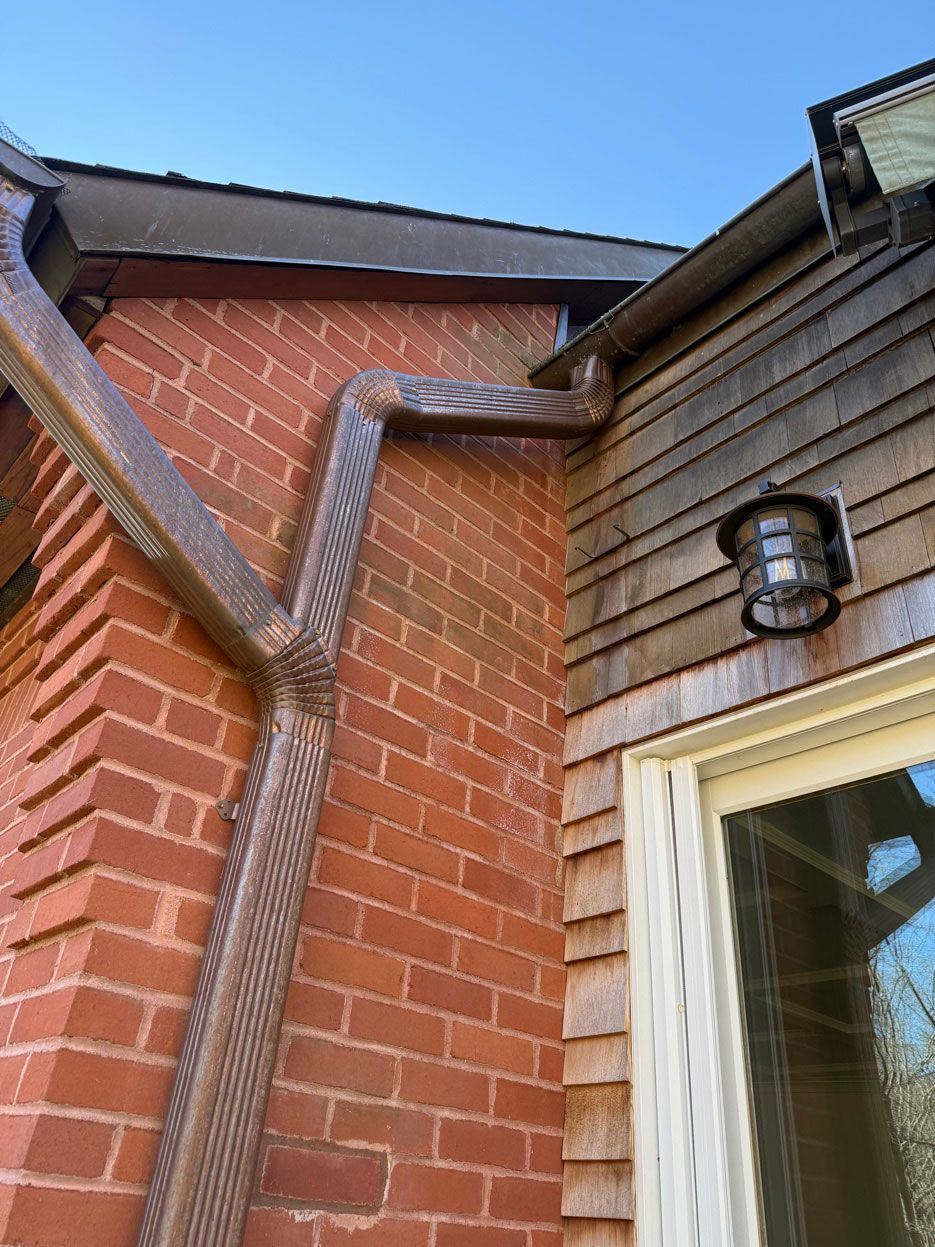 A copper downspout runs along a red brick wall adjacent to gray wood siding and a window.