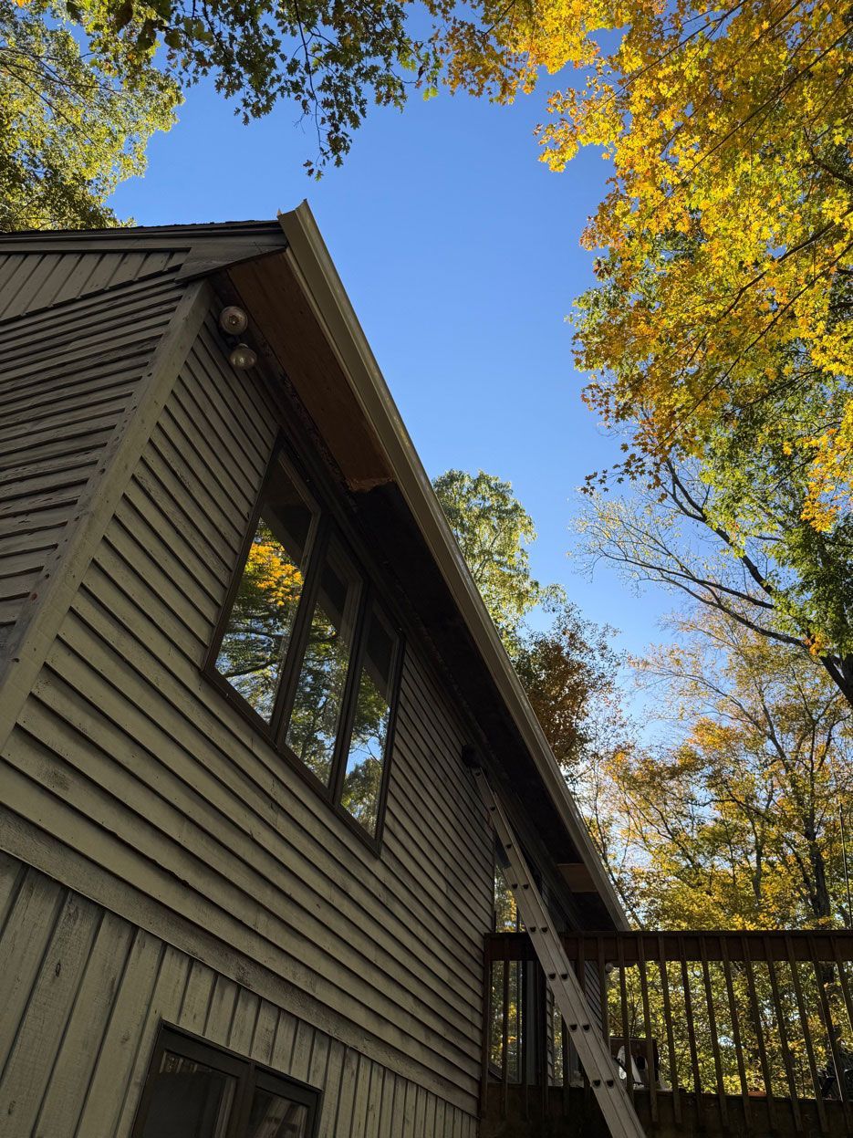 A low-angle view of a house with beige siding, a window, and a ladder, set against autumn trees and a clear blue sky.