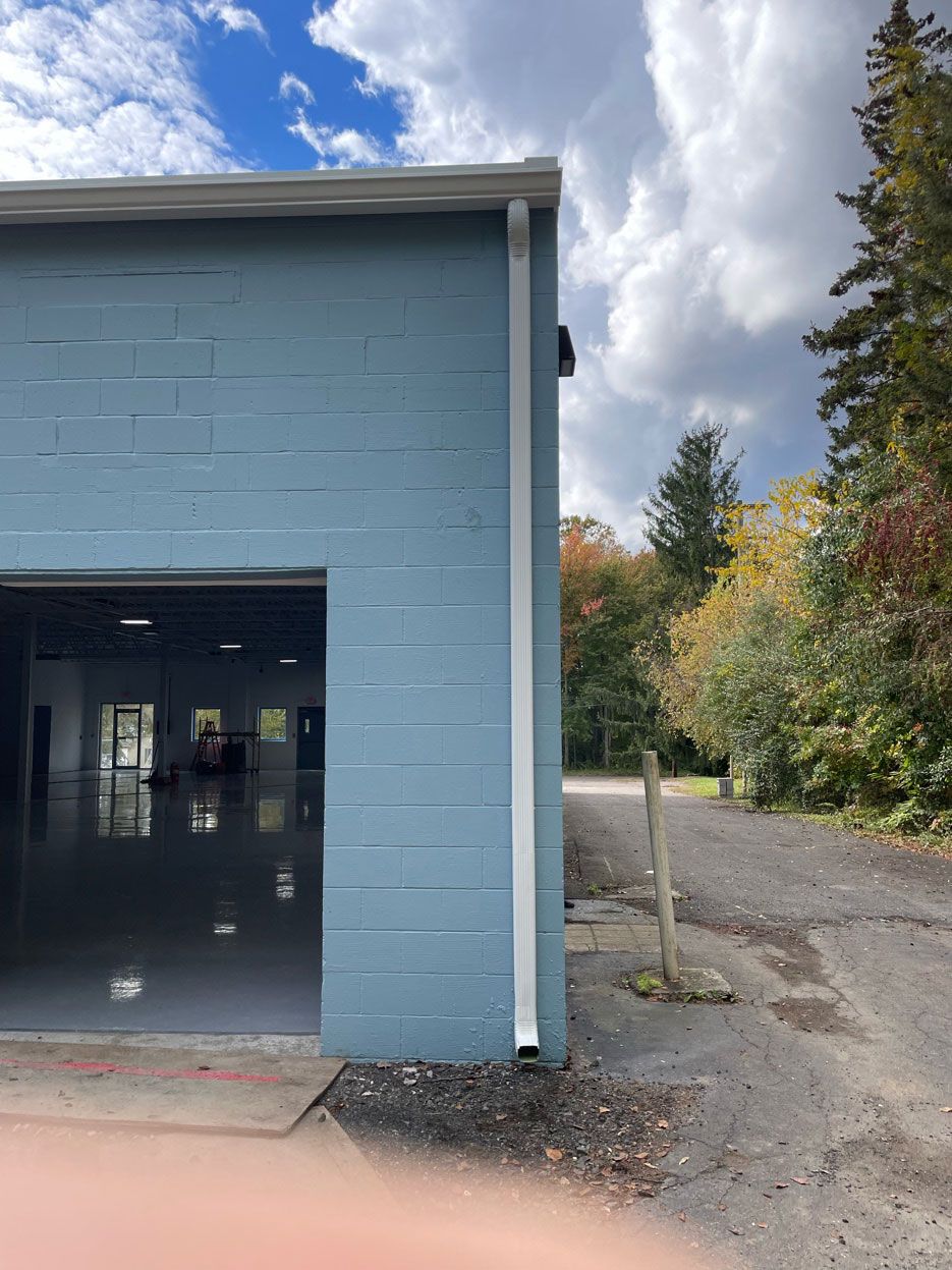 Light blue concrete block exterior of a building with a white downspout on the corner, overlooking an outdoor gravel area.