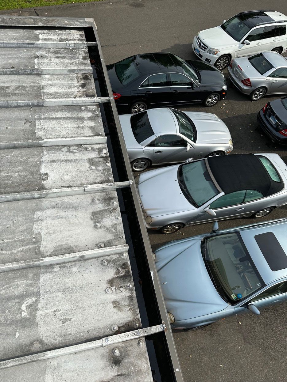 A high-angle view of a weathered concrete roof edge bordering a parking lot filled with several silver and dark cars.