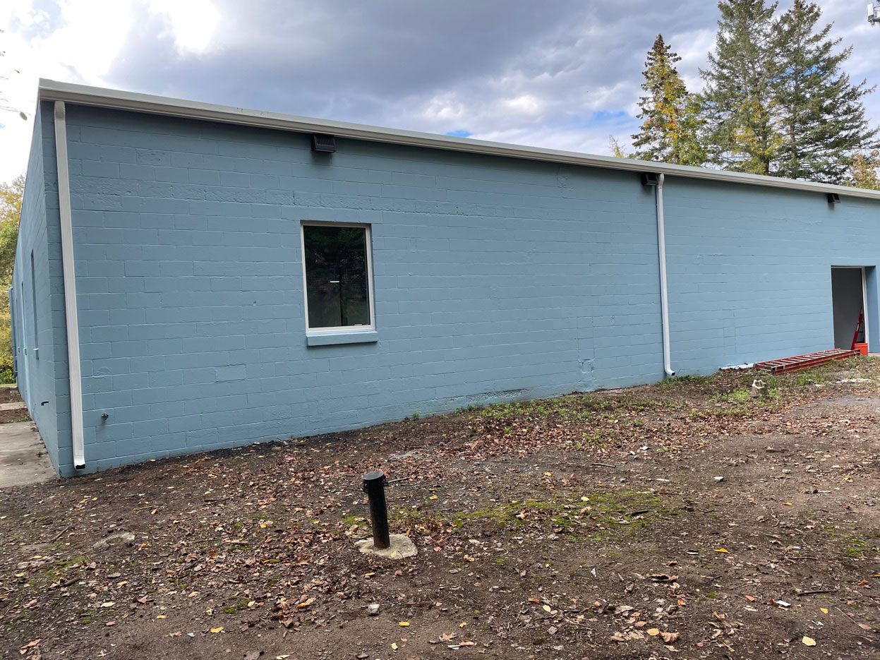 Light blue, single-story cinder block building with a single window and an open doorway on a dirt lot.
