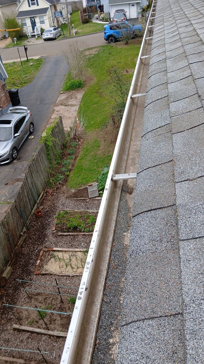 A high-angle view of a gray shingled roof and a white rain gutter, with a residential street visible below.