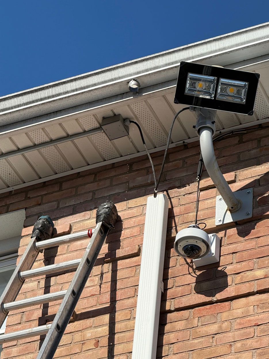 An outdoor wall-mounted security camera and LED floodlight installed on a brick wall with an extension ladder nearby.