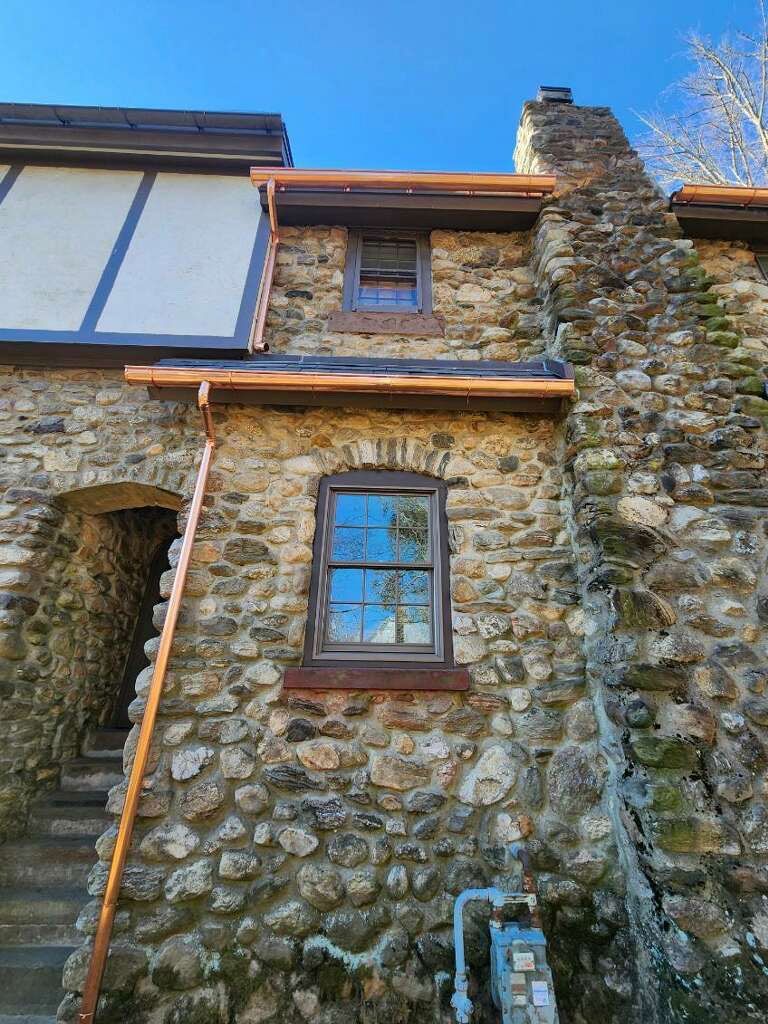 A stone-walled house exterior with new copper gutters installed beneath a dark wood overhang and a chimney on the right.
