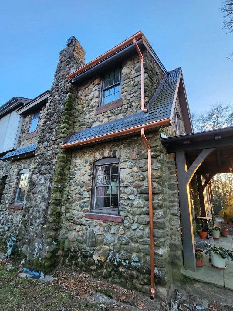 A stone house exterior with a prominent chimney, dark roof tiles, and new copper gutters set against a clear blue sky.