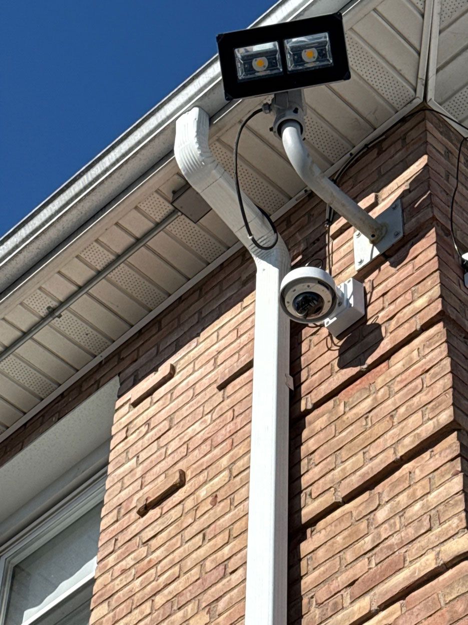 A security camera and rectangular LED floodlight are mounted to the exterior corner of a brick building.