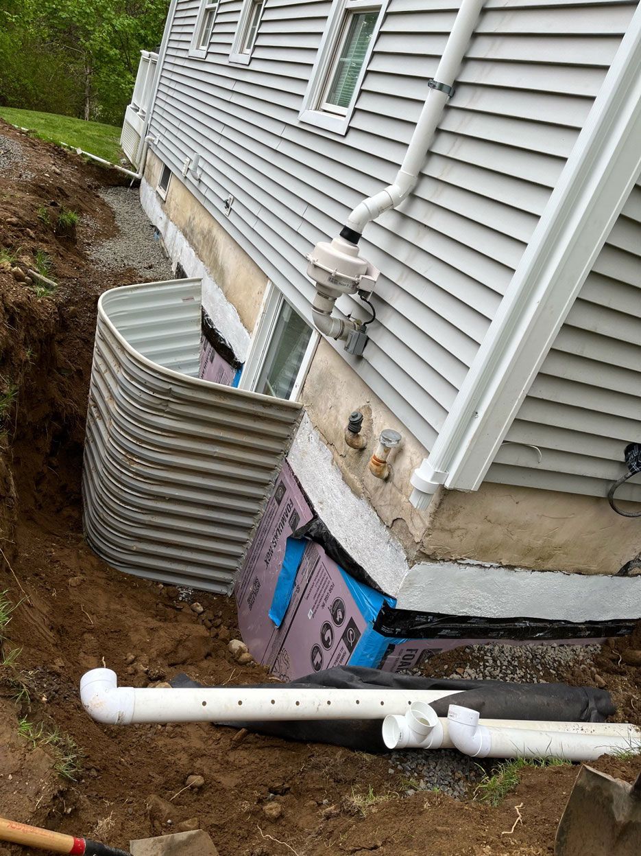 A side view of a house foundation under repair, showing a window well, foam insulation, and perforated drainage pipes.