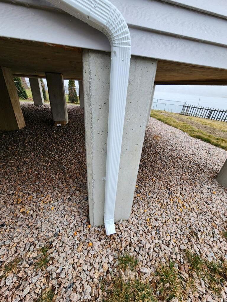 A white gutter downspout attached to a concrete pillar under a building, with gravel ground and a water view.