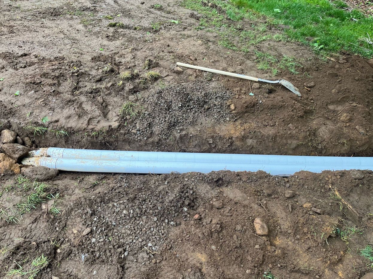Two light blue corrugated pipes lie side-by-side in a narrow dirt trench, with a shovel resting on the soil nearby.