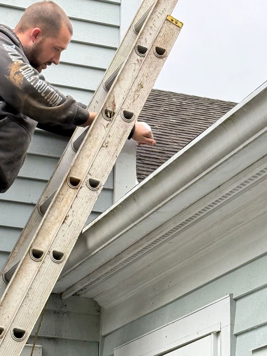 A person on a ladder reaching up toward the roofline of a light blue house to inspect or repair the gutters.