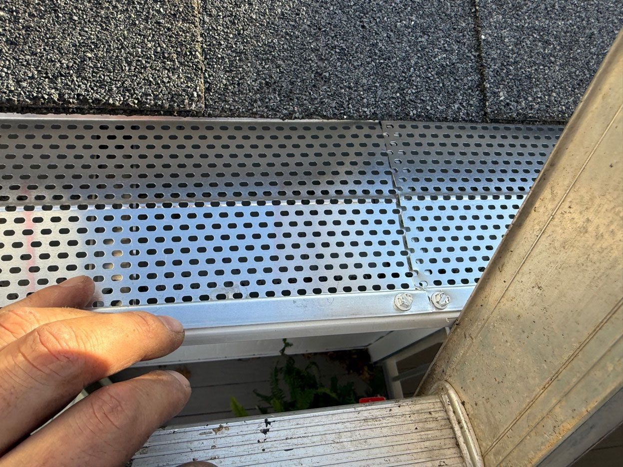 A hand holds a metal, perforated gutter guard in place on top of a rain gutter installed under a shingled roof.