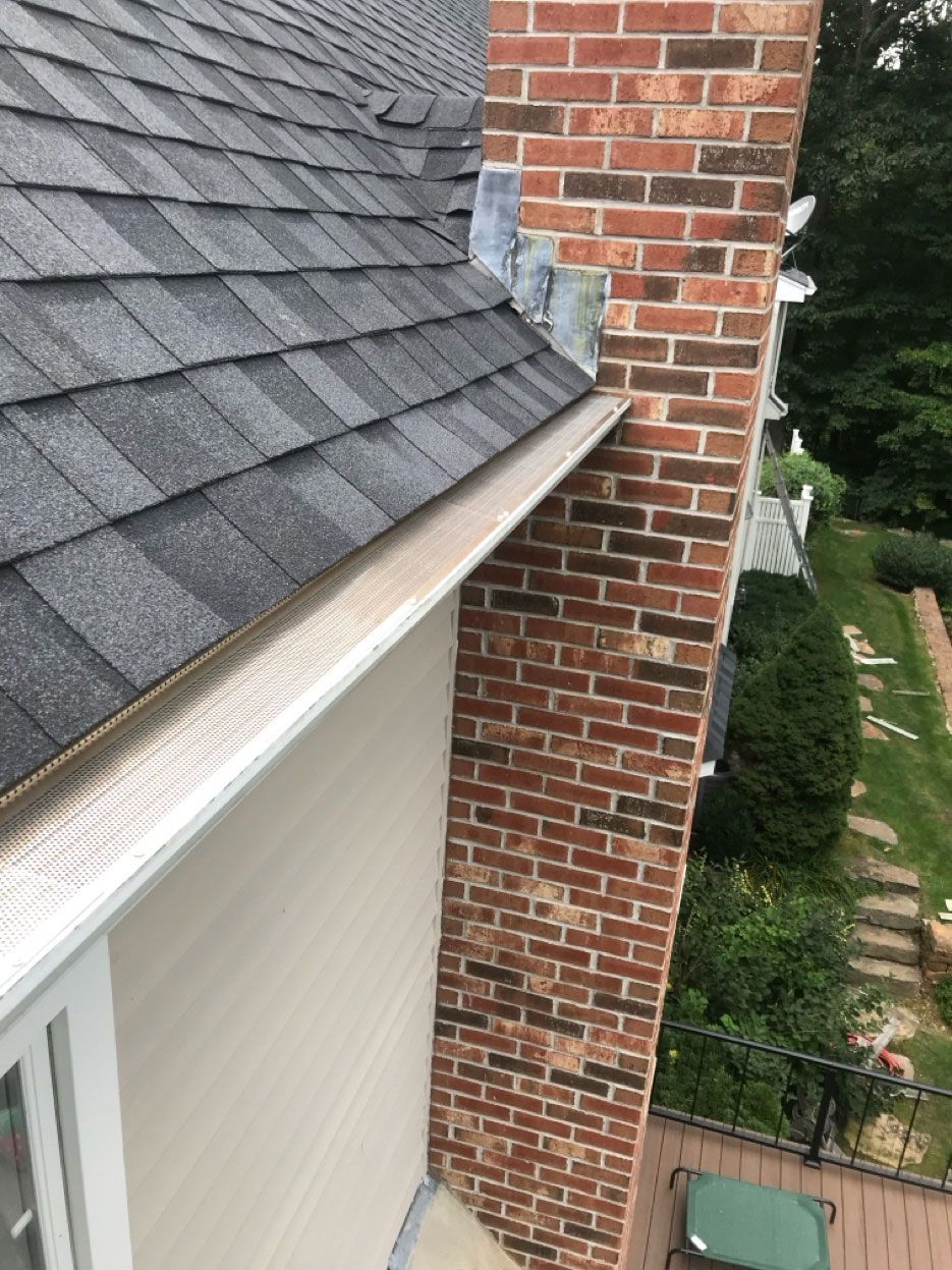 An elevated view of a house roofline, featuring dark shingles, a brick chimney, and a gutter with a protective screen.