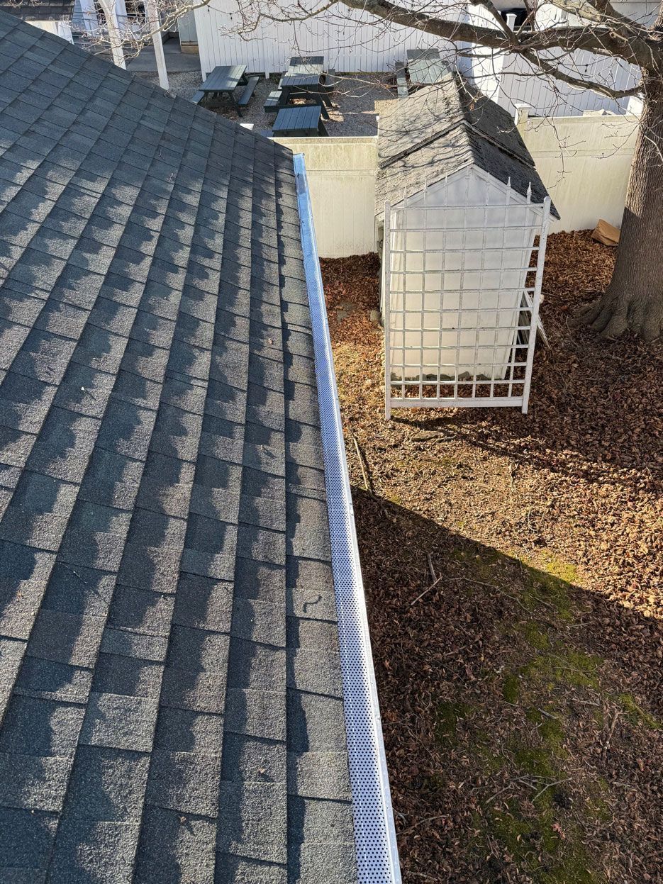 A high-angle view of a gray asphalt shingle roof edge with a white gutter guard, overlooking a yard with a garden trellis.