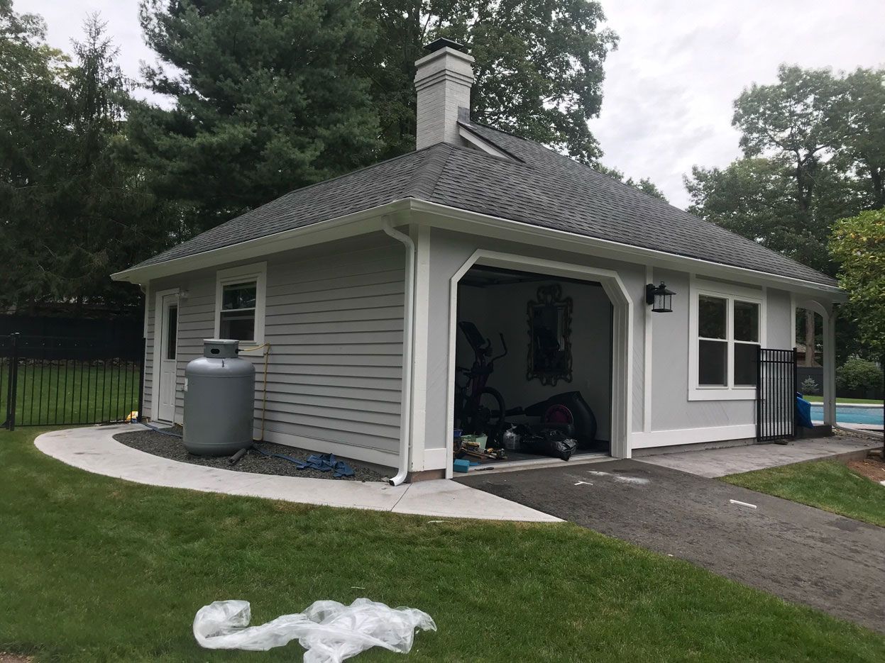 A gray detached garage with a white chimney, a propane tank, and an open bay door, set on a lawn with a paved path.