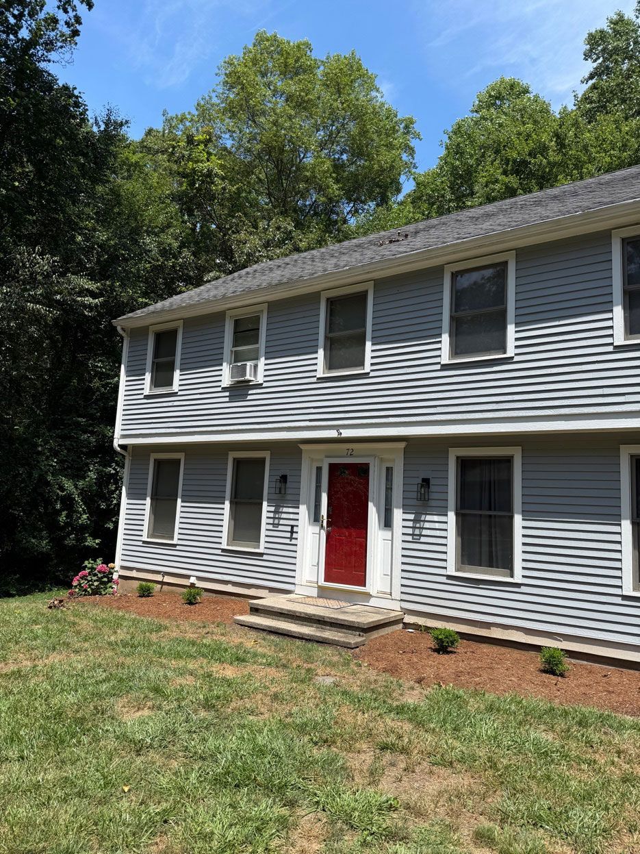 A two-story house with gray wood siding, a bright red front door, and a brown mulch yard, surrounded by green trees.