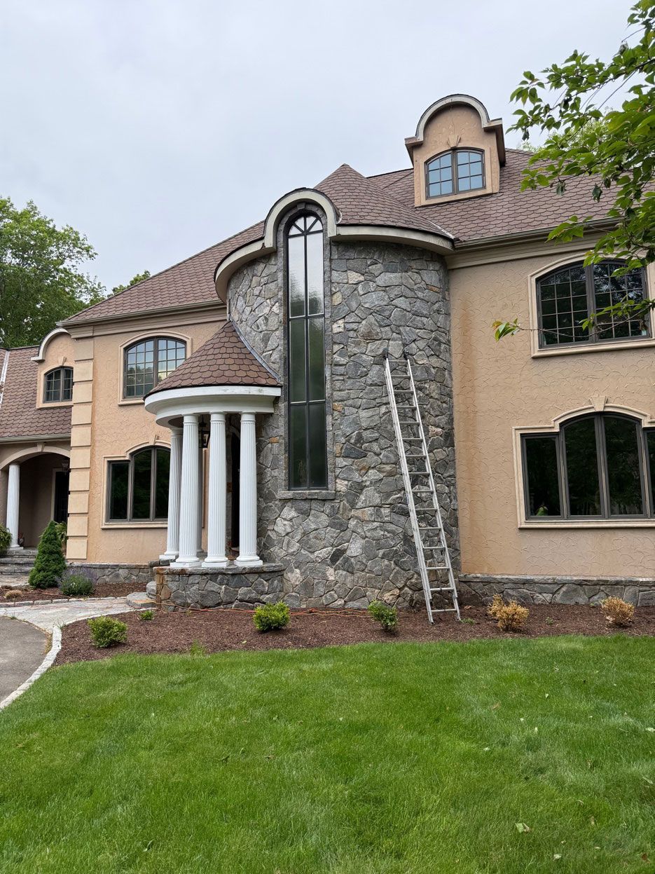 A two-story stucco and stone house with a curved tower section, an arched entrance with columns, and an extension ladder.