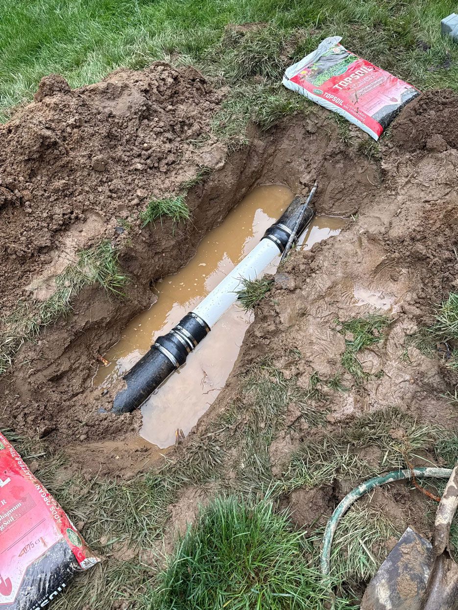A trench in a grassy lawn reveals a PVC pipe connected to existing black pipe using two rubber couplings.