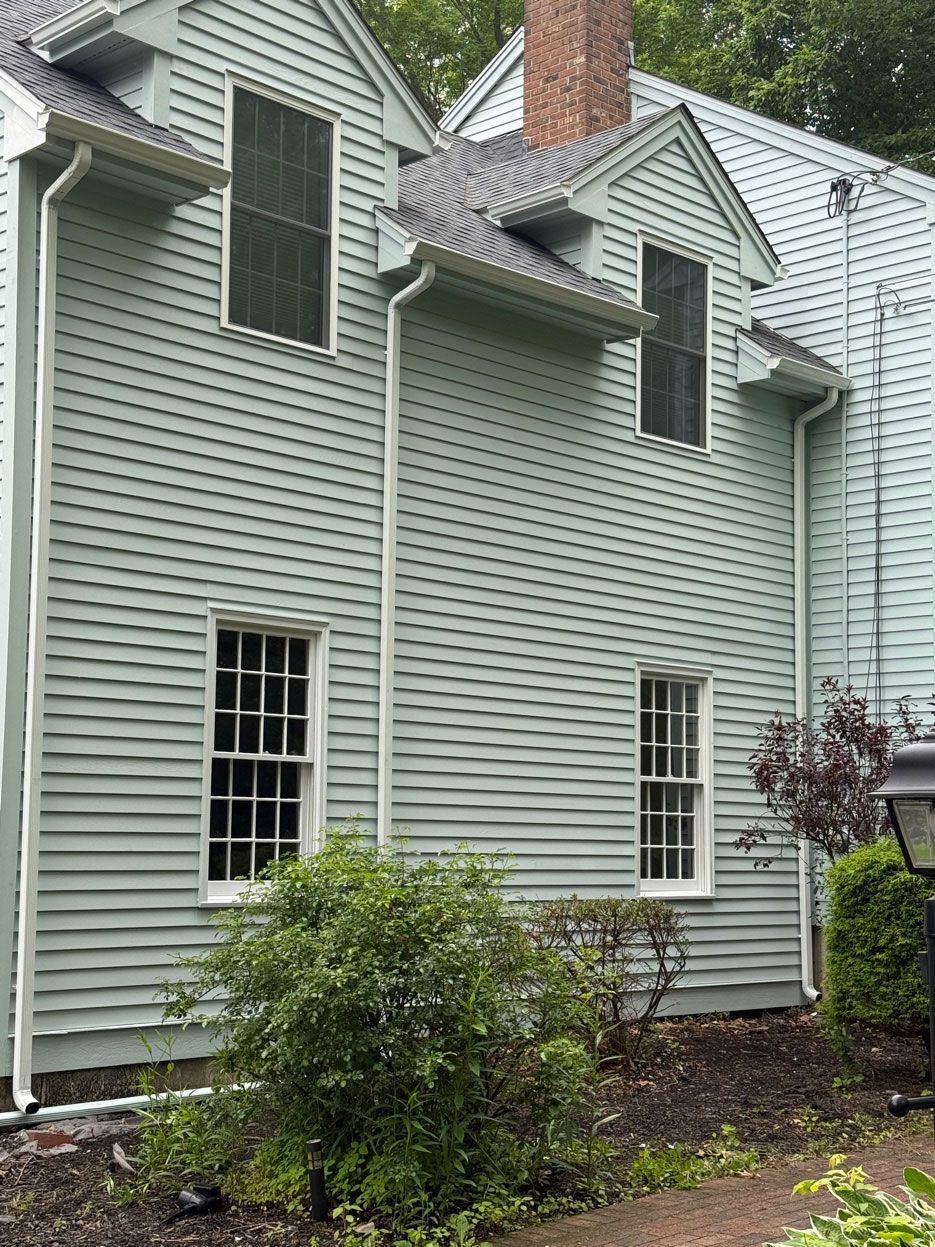 A two-story light green house exterior with white trim, dormer windows, and a brick chimney.