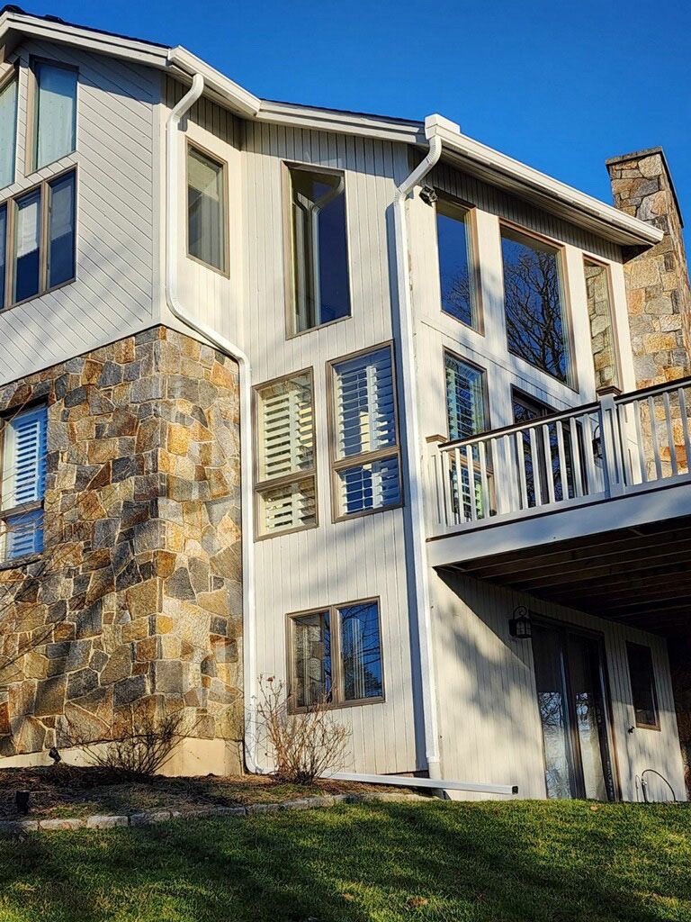 Two-story house exterior with stone and light siding, featuring large windows, a deck, and a green lawn under a clear sky.